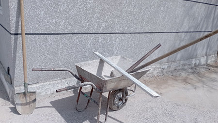 construction wheelbarrow-trolley rake, pitchfork inside. Shovel stands against the wall next to the wheelbarrowの写真素材