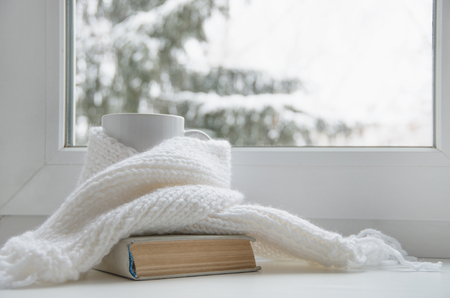 Mug of hot tea and warm woolen knitting on windowsill against snow landscape from outside.の写真素材