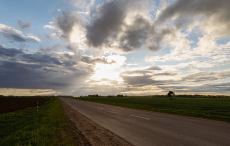Sunset over the road between fields and meadows. Beautiful clouds. Horizontal photo.の写真素材