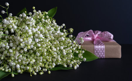 Studio photo. Greeting card. A bouquet of fragrant white lilies of the valley on a dark background with a gift in craft packaging with a pink ribbon.の写真素材
