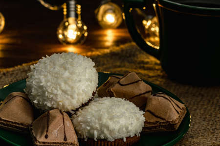 Close-up of souffle cakes with coconut flakes and crispy biscuits on a saucer with a cup of coffee or tea. Photo in a dark key, warm subdued light from the lights of a lamp garland. green tea set. selective focus.の写真素材
