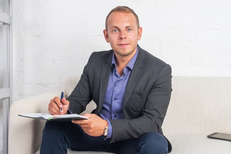Handsome businessman sitting on couch with notebook at home in the living room, looking cameraの写真素材