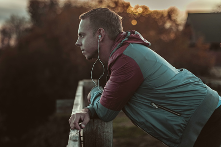 Attractive young athletic man standing on the beach and looks into the distance of the river to the bridge, listening to musicの写真素材