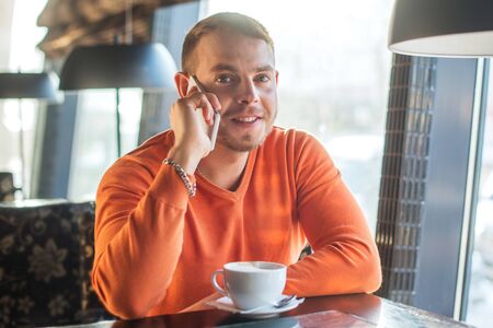 Handsome young man working, talking on the phone and smiling while enjoying coffee in cafe, looking...