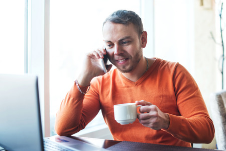 Handsome young man working with notebook, talking on the phone, smiling,  while enjoying coffee in cafe.の写真素材