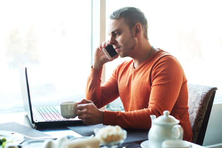 Handsome young man working with notebook, talking on the phone,, while enjoying coffee in cafe.の写真素材