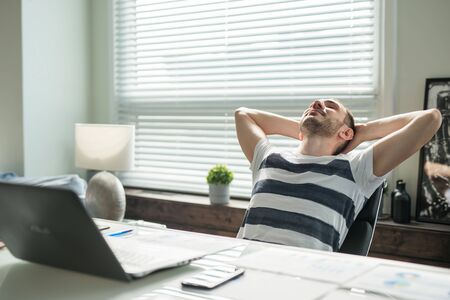 Young man businessman sitting in an office at a table with a laptop, put his hands behind his head, closed his eyes and rests.の写真素材