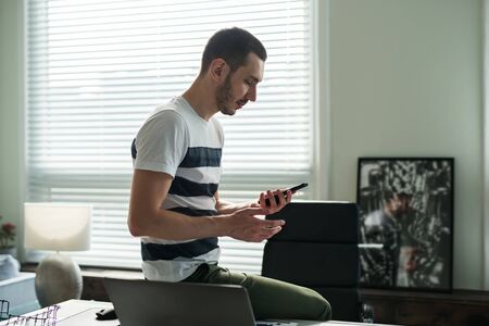 Young man businessman stands next to a work desk and looks at his phone in the office.の写真素材