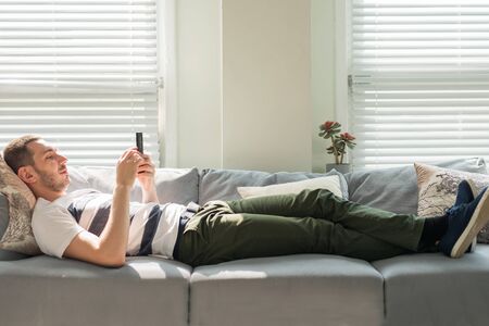Handsome young man looking his smart phone while, lying on the couch at home.の写真素材