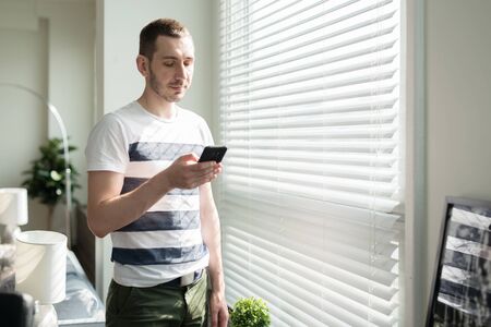 Young man businessman is standing near the window, looking at the phone in office.の写真素材