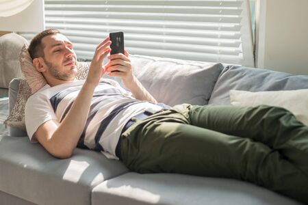 Handsome young man looking his smart phone while, lying on the couch at home.の写真素材