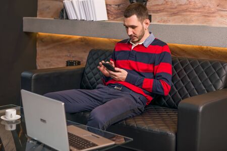 Young man working on a laptop at his desk in the office sitting on a sofa and holding a phone.の写真素材