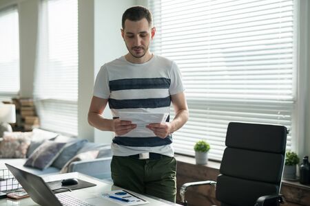 Young business man working with laptop at his desk in the office holds documents.の写真素材