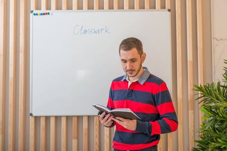 Young man standing at the blackboard and looking at a notebook.の写真素材