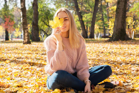 Young pretty woman is sitting on ground in autumn park with leaves in her hands.の写真素材