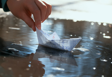 childrens hand of the schoolboy launches a small paper boat made of a school notebook in a spring early puddle on a Sunny bright dayの写真素材