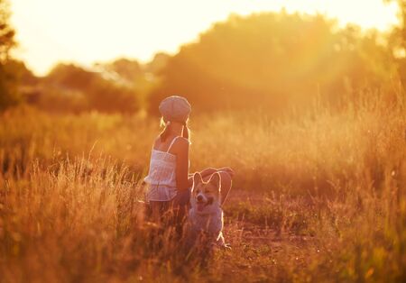 girl in a cap and a cute red puppy of corgi sit on an old suitcase not off the road into a field and waiting for transport when traveling to sunny summer eveningの写真素材