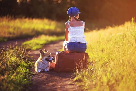 girl in a cap and a cute red puppy of corgi sit on an old suitcase not off the road into a field and waiting for transport when traveling in the summer sunny dayの写真素材