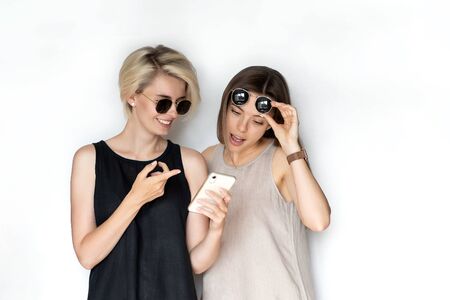Two beautiful young girls in summer dresses look at the phone and smile on the white background. High quality photoの写真素材