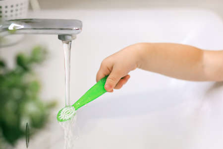 The babys hand holds a tooth brush under a stream of water close-up at home in the bathroom, concept: childrens hygiene.の写真素材