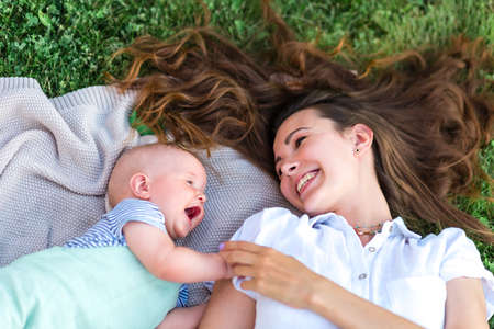 Cute baby and his mom look at each other and laugh, they lie on their backs on the lawn grass and blanket. The concept of family relations.の写真素材