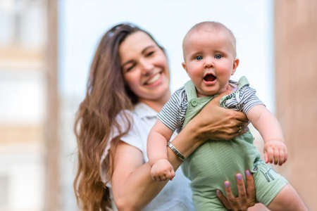 A portrait cute baby looking straight at the camera on the mother hands, mom blurry on a urban background in the summer dayの写真素材