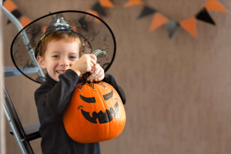 A CHEERFUL AND CUTE BOY OF 5 YEARS OLD HOLDING AN ORANGE PUMPKIN WITH A PAINTED GRIMACE FOR HALLOWEEN, INDOORS WITH COPYSPACE.の写真素材