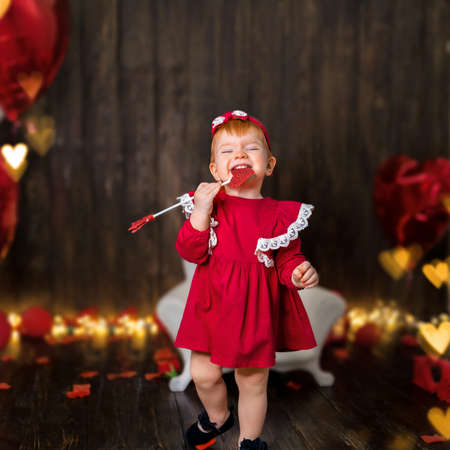 a one-year-old girl with a happy emotion in a red dress with a cupid arrow on a dark wooden background with red hearts and bokeh.の写真素材