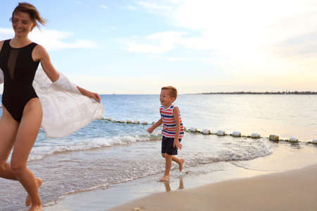 A young mother in a swimsuit and a charming laughing son 5 years old play and run along the sandy beach at sunset, the concept of Active family summer vacation at sea.の写真素材
