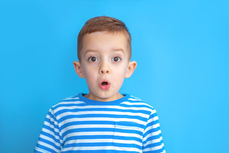 Close-up portrait of surprised Caucasian boy in striped T-shirt with brown eyes and white skin on blue studio background. Cute redhead boy looking totally shocked, opening mouth in surprise.の写真素材