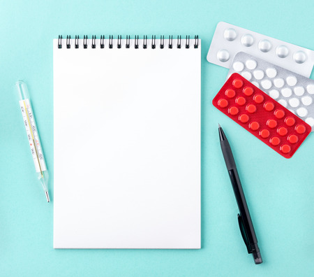 open notebook with blank white clean page to write the treatment plan of the disease. Pills, pen, paper, thermometer on blue background, top view.の写真素材