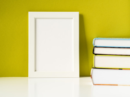 Blank white frame and stack of books on a white table against the olive colored wall with copy space