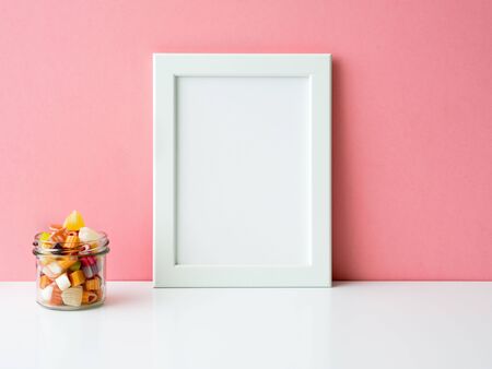Blank white frame and lollipops in jar on a white table against the pink wall with copy spaceの写真素材