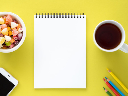 Open notepad on the spiral with a clean white page, a Cup with tea, caramels in a bowl, a smartphone and color pencil on a bright yellow background colorの写真素材