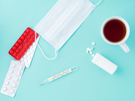 Pills, thermometer, medical face mask, tea in a mug on a blue background, top view. First aid, care...