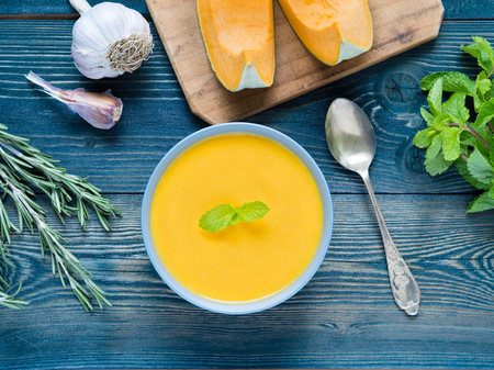Creamy soup of pumpkin on dark blue wooden background, rosemary, garlic, mint, slices of pumpkin. Top view.の写真素材