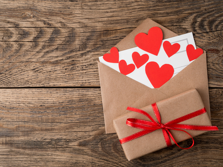 card and red hearts in open envelope from brown Kraft paper. Gift box with red ribbon on wooden aged...