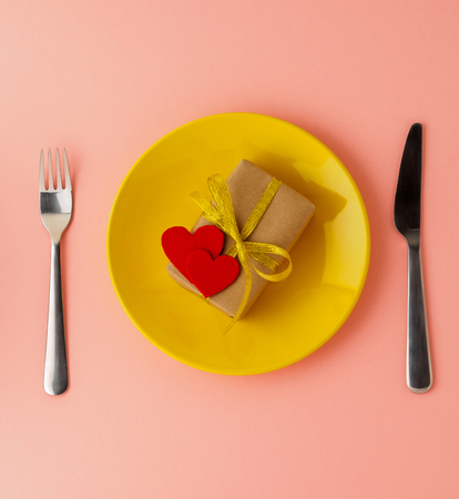 gift, pack of Kraft brown paper with a gold ribbon, two felt hearts on a yellow plate, knife, fork on a bright pink background, top view, copy space for text. Valentine's day.の写真素材