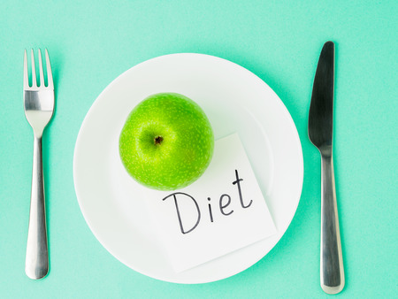 Ripe raw juicy green Apple, slip of paper with inscription diet on a white plate, fork, knife on a bright turquoise blue colour table, top view. Diet as lifestyle.の写真素材