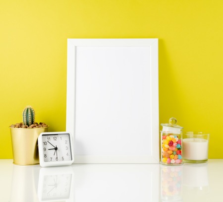 Blank white frame, clock, succulent, candy on a white table against the yellow wall. Mockup with copy space.の写真素材