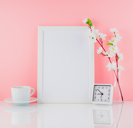 Blank white frame, flower, clock and cup of coffee or tea on a white table against the pink wall...