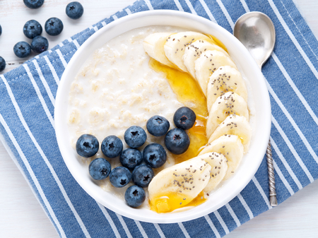 Oatmeal with bananas, blueberries, chia, jam, honey, blue napkin on a white wooden background. Close-up. Top viewの写真素材