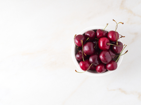 The red dark sweet cherries in white bowl on stone white table, top view, copy space