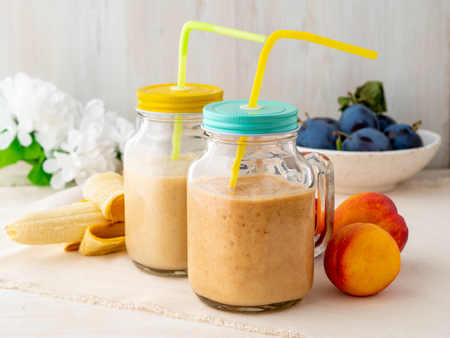 banana peach nectarine smoothie in two jars, fresh fruit milkshakes on the white rustic wooden table, side viewの写真素材