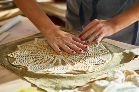 Woman making pattern on ceramic plate, hands close-up, focus on palms with clay and lace. Creative...