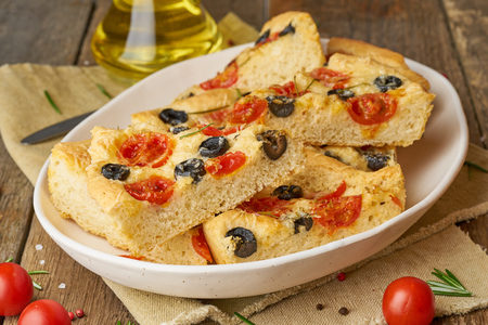 Focaccia, pizza in plate with tomatoes, olives and rosemary. Chopped Italian flat bread. Side view, a wooden tableの写真素材