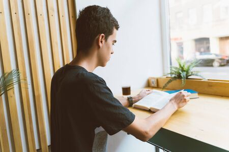 Young boy with black hair sitting by the window in the library reading a big book, teenager in casual clothes in libraryの写真素材