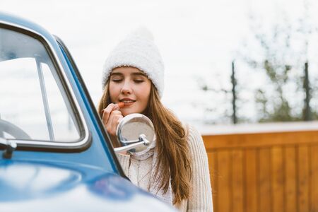A beautiful young girl looks in the car mirror and paints her lips. Woman with long dark thick hair next to a blue retro car in autumn or winter, copy spaceの写真素材