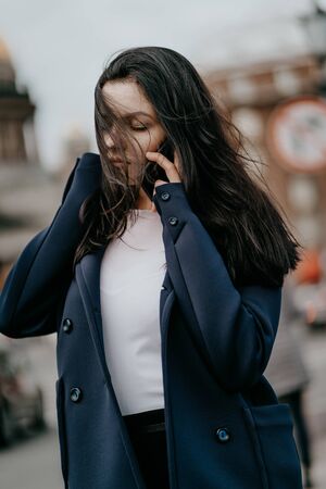 Woman with long hair talking on phone. Autumn or winter, girl in outdoor. Beautiful intelligent brunette in street of Saint-Petersburg in city center, copy space, verticalの写真素材