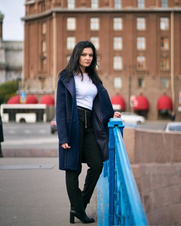 Charming thoughtful fashionably dressed woman with long dark hair travels through Europe, standing in city center of St. Petersburg. A beautiful girl wanders alone through autumn streets, verticalの写真素材
